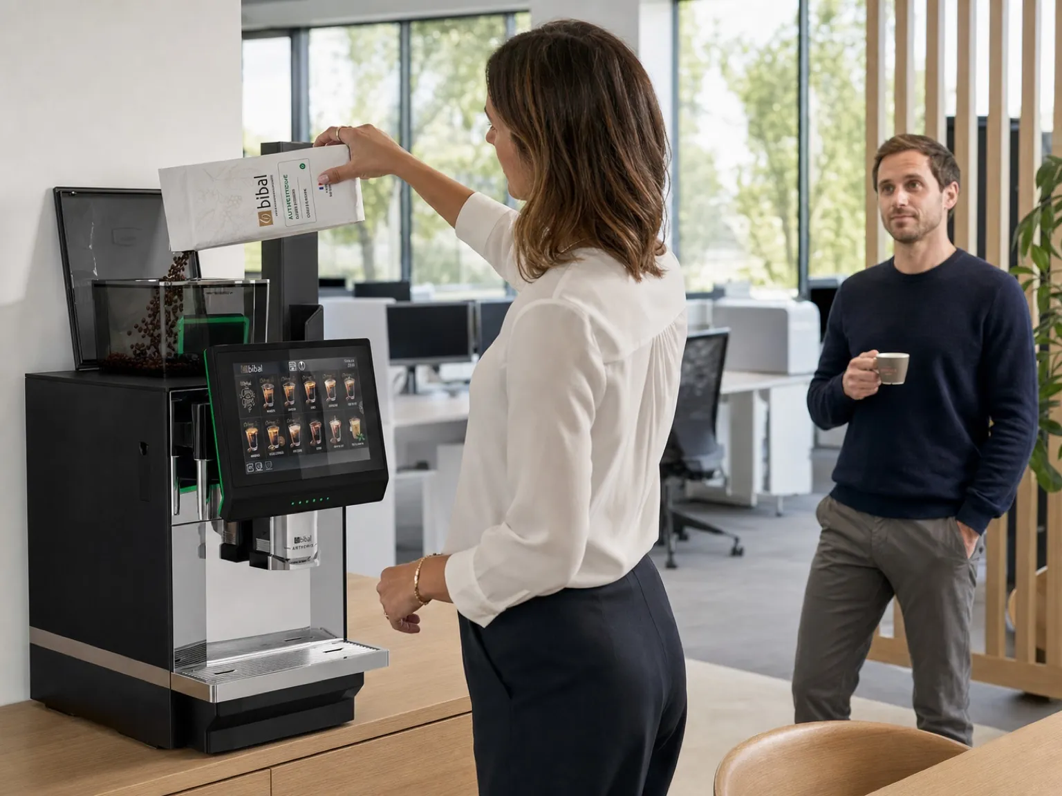 Open space d'une PME contemporaine : espace café sur un meuble en chêne clair, l'office manager remplit le bac à grains de la machine pendant qu'un collègue prend sa pause