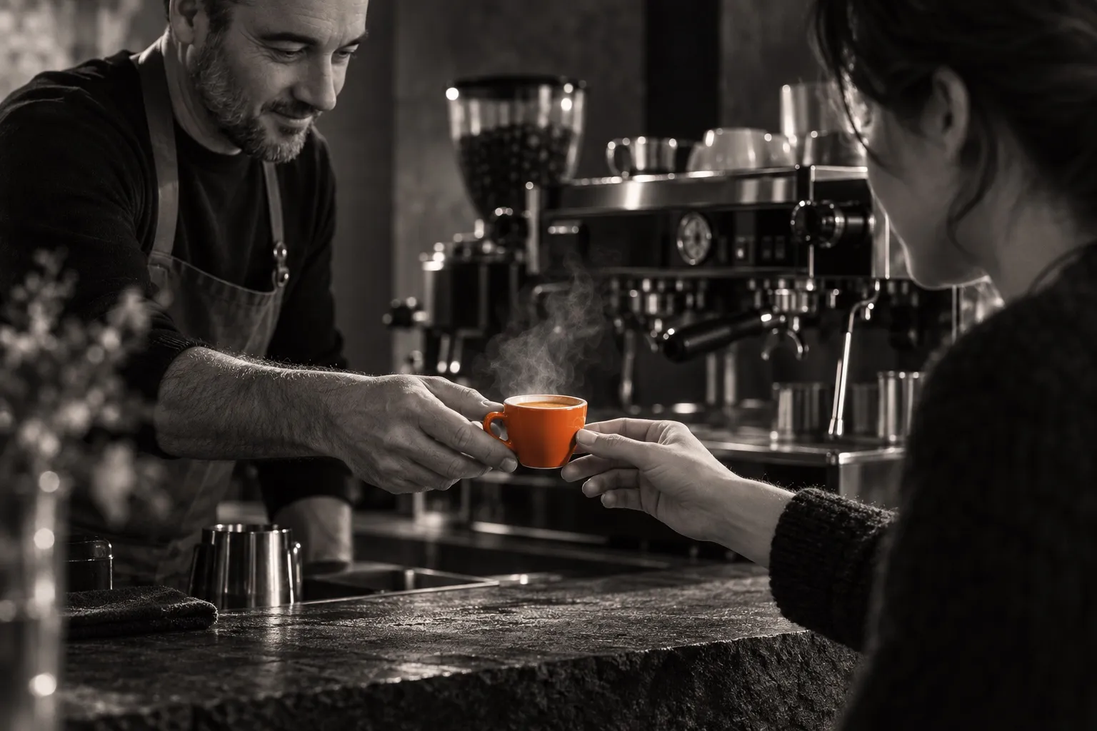 Dans la vapeur d'un coffee shop, un barista tend une tasse espresso orange à une cliente devant la machine — photographie en noir et blanc chaud, la tasse pour seule couleur